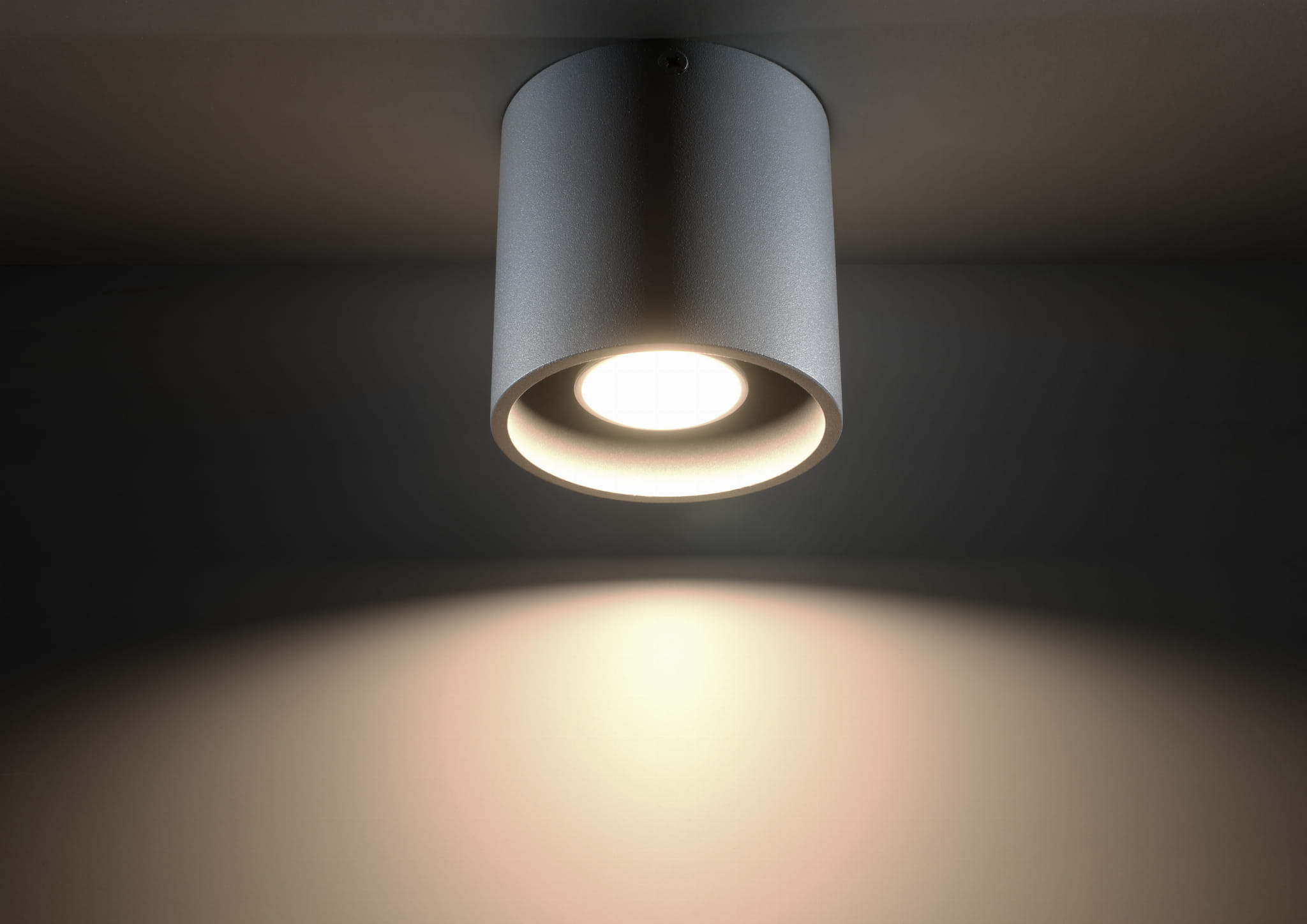 Lampa sufitowa Orbis 1-punktowa w kolorze szarym, o cylindrycznym kształcie. Widoczny klosz z otworem na żarówkę, emitujący światło w dół. Ujęcie z dołu, na jednolitym tle, podkreślające efekt świetlny na ścianie.
