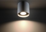 Lampa sufitowa Orbis 1-punktowa w kolorze szarym, o cylindrycznym kształcie. Widoczny klosz z otworem na żarówkę, emitujący światło w dół. Ujęcie z dołu, na jednolitym tle, podkreślające efekt świetlny na ścianie.
