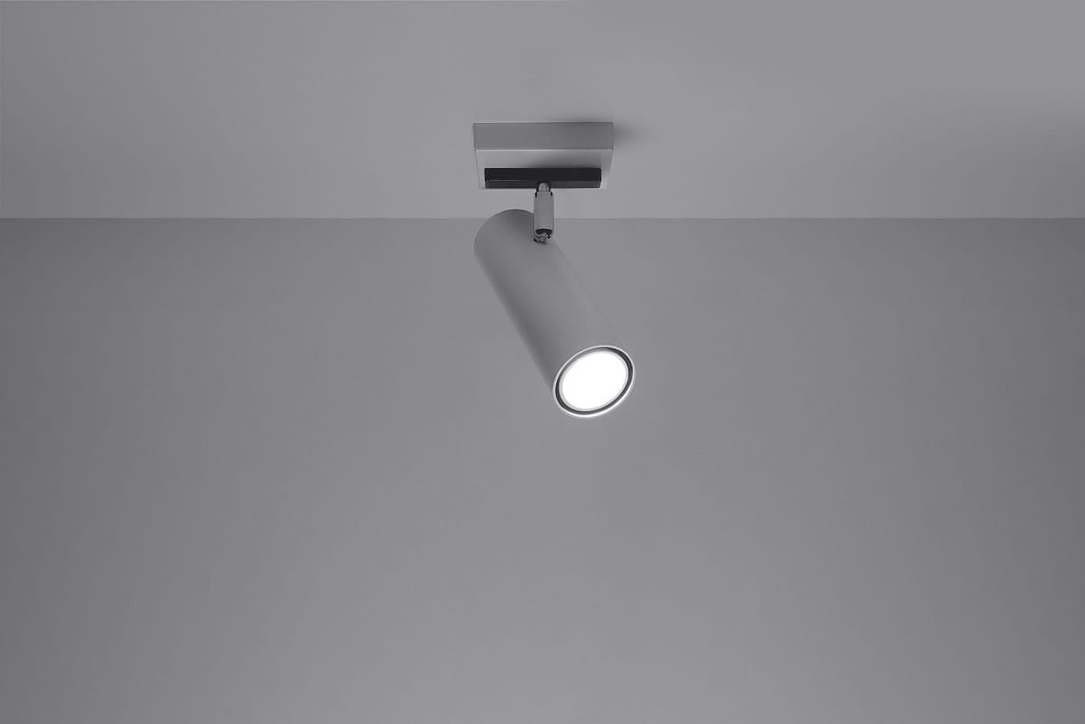 Biała lampa sufitowa (spot) Direzione, 1-punktowa, z cylindrycznym kloszem. Ujęcie z dołu, lampa zamontowana na suficie, emitująca światło. Prosty design, harmonizujący z minimalistycznym wnętrzem. Tło szare, bez dodatkowych elementów.