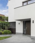 Czarny kinkiet elewacyjny 2-punktowy Exe 8330 zamontowany obok drzwi wejściowych w nowoczesnym budynku. Produkt ma prosty, cylindryczny kształt i jest umieszczony na jasnej, gładkiej ścianie. W tle widoczne są krzewy i trawniki.