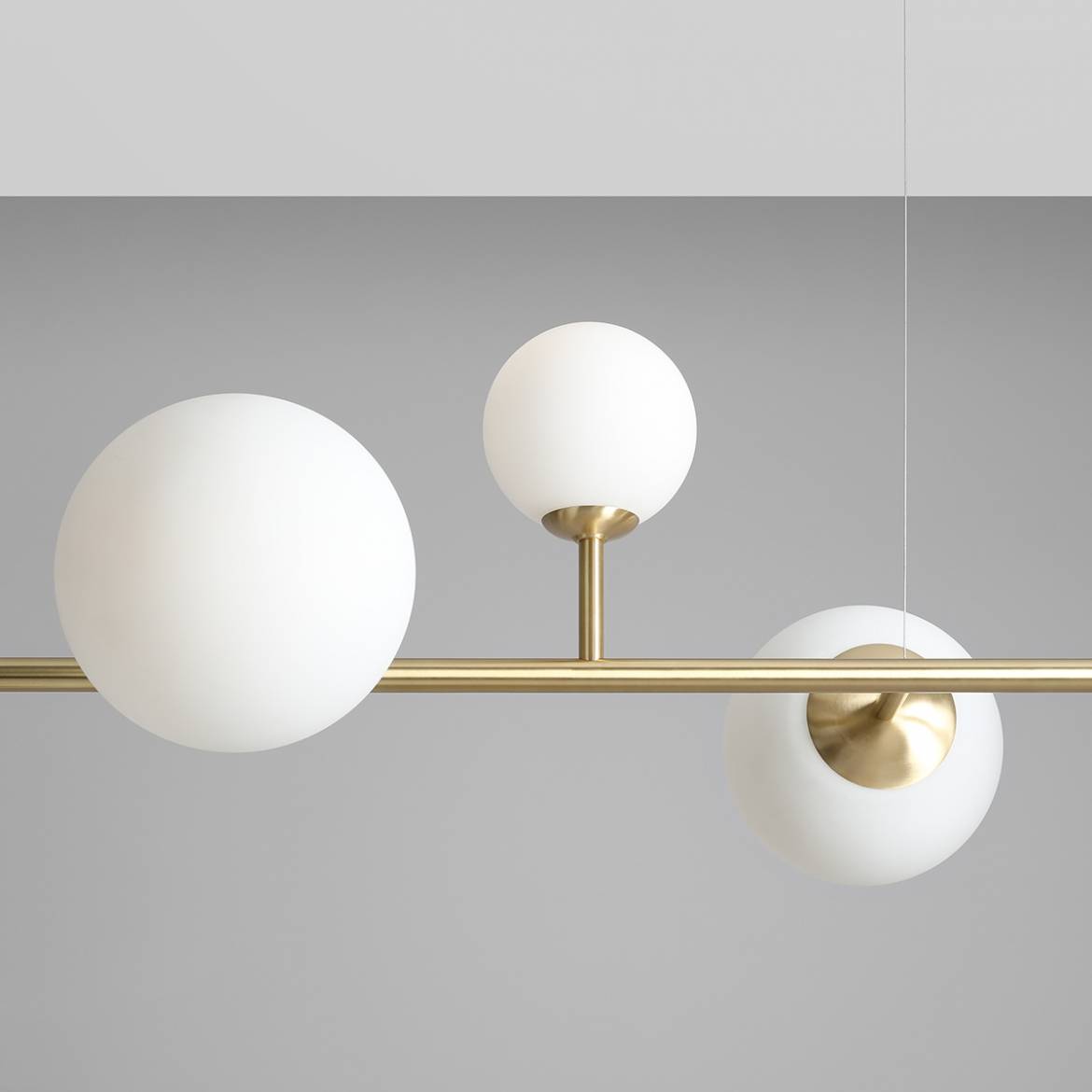 Lampa wisząca Dione, 6-punktowa, w kolorze mosiądzu. Posiada trzy białe, okrągłe klosze z matowego szkła zamocowane na prostym, poziomym ramieniu. Ujęcie z góry, na tle szarym, ukazujące detale konstrukcji.