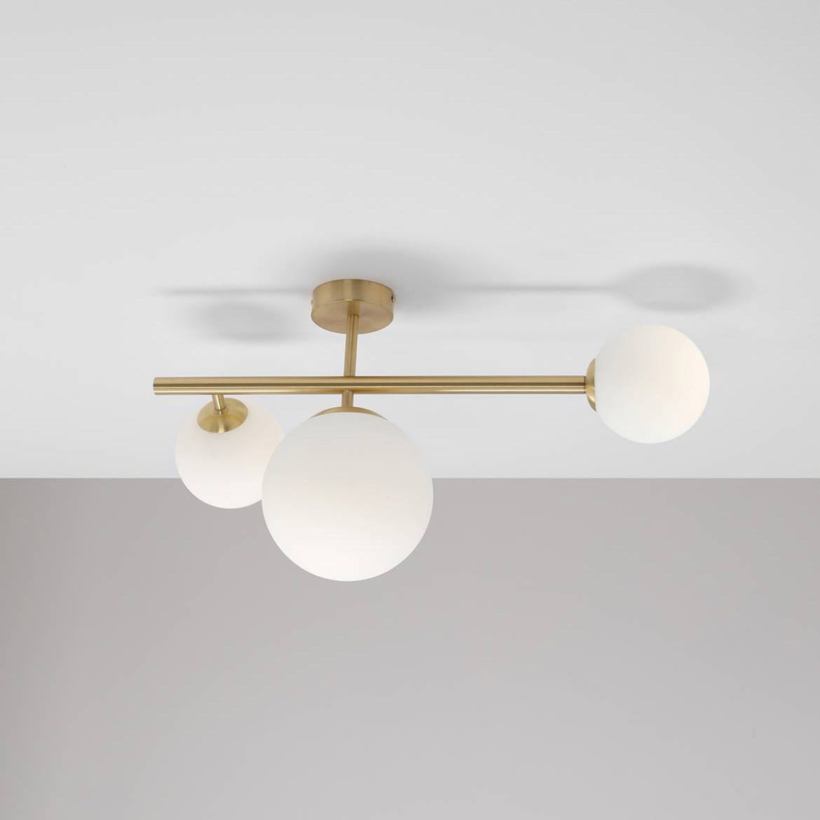 Lampa sufitowa Dione 3-punktowa w kolorze mosiądzu, z trzema okrągłymi kloszami z białego szkła. Ujęcie z góry, pokazujące całość lampy oraz jej mocowanie do sufitu. W tle jasnoszare ściany.