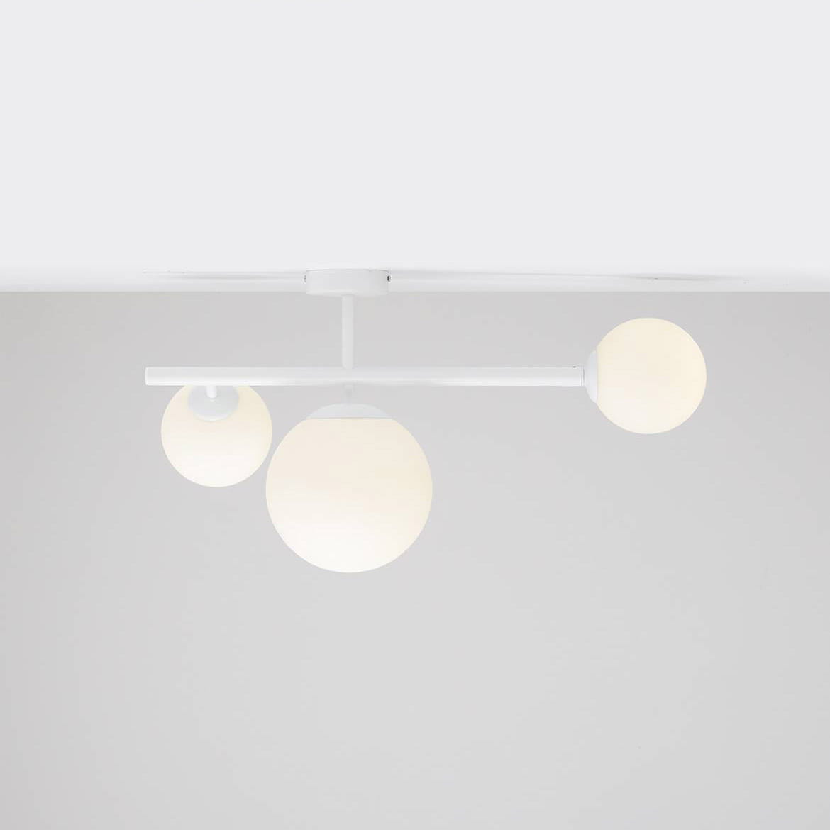 Lampa sufitowa Dione 3-punktowa w kolorze białym, z trzema okrągłymi kloszami w odcieniach bieli. Klosze zawieszone na prostym, poziomym ramieniu. Ujęcie z dołu, ukazujące cały produkt na jednolitym tle.
