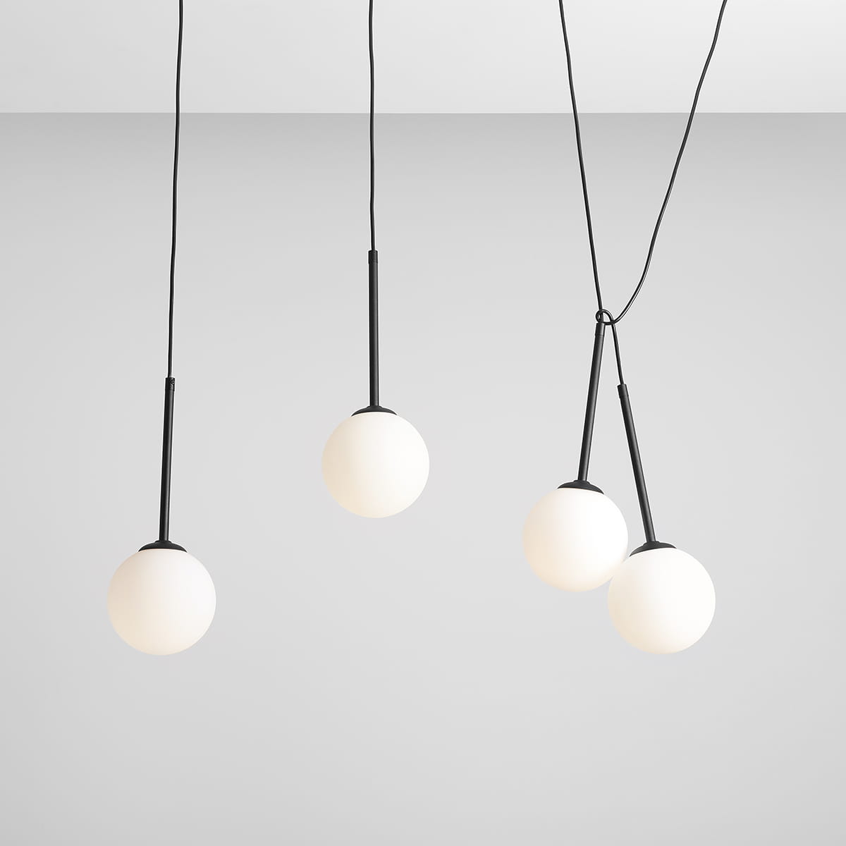 Czarna lampa wisząca Bosso z czterema punktami świetlnymi. Każdy z kloszy w kształcie kuli wykonany z matowego szkła, zawieszony na cienkich, czarnych linkach. Lampa wisi w prostej aranżacji na jasnym tle.