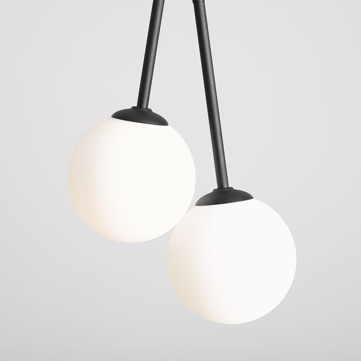 Czarna lampa wisząca Bosso z czterema punktami świetlnymi. Dwie białe, okrągłe klosze z matowego szkła zawieszone na czarnych prętach. Ujęcie z bliska, ukazujące detale mocowania kloszy. Tło w jasnym odcieniu.