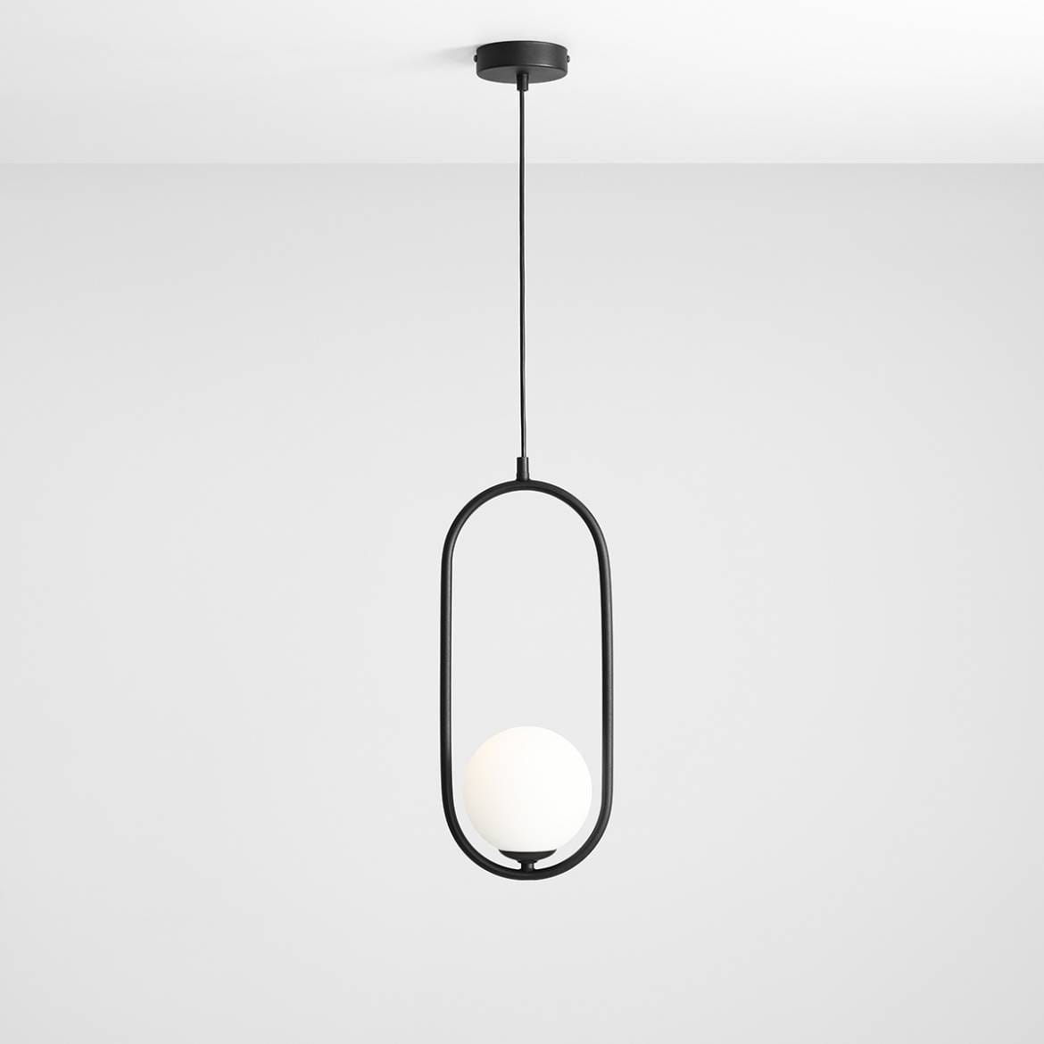 Lampa wisząca Riva 1-punktowa w kolorze czarnym, z okrągłym, białym kloszem. Produkt zawieszony na cienkim przewodzie, w minimalistycznej, owalnej ramie, montowany do sufitu. Tło jasnoszare, podkreślające nowoczesny design.