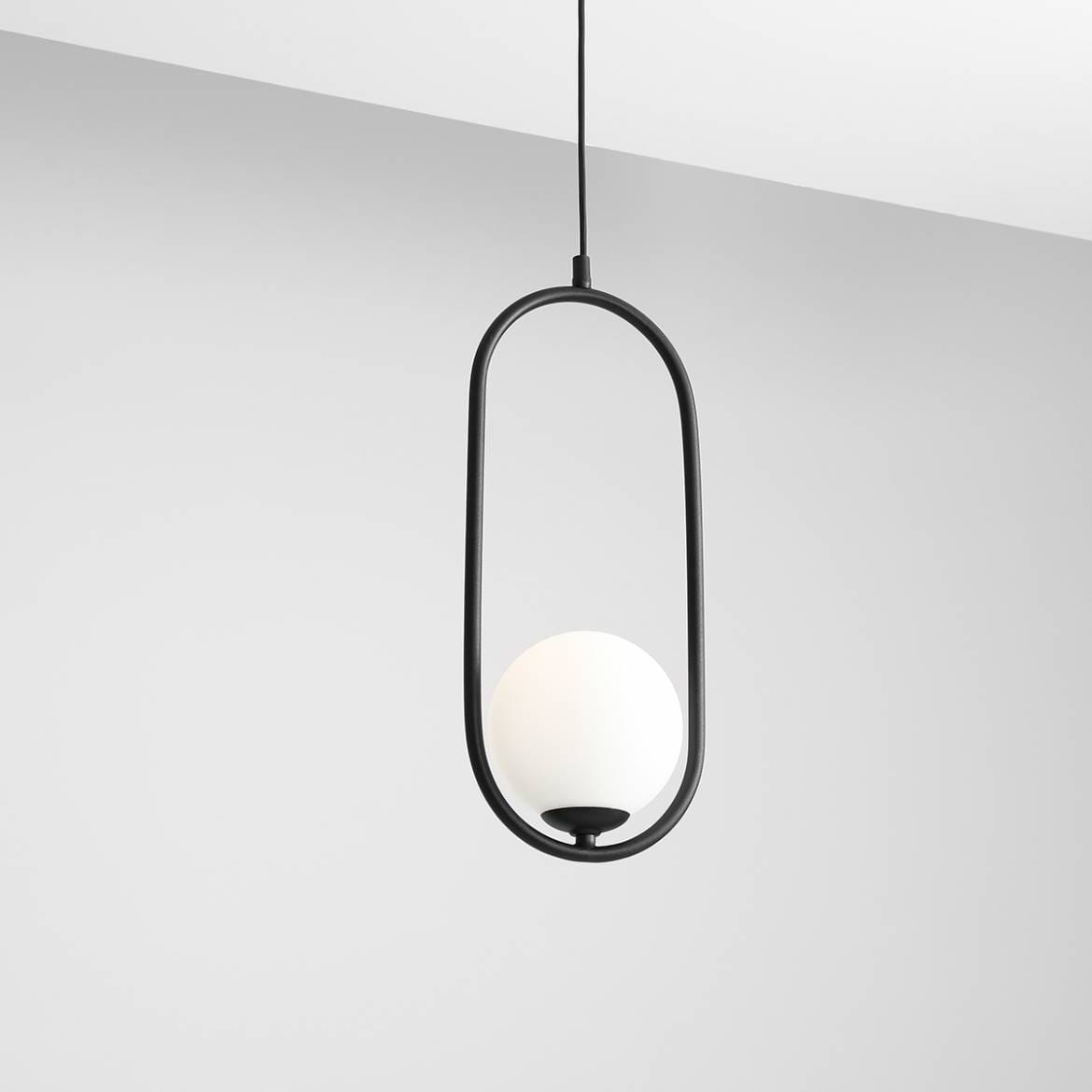 Czarna lampa wisząca Riva 1-punktowa z okrągłym, białym kloszem umieszczona w owalnej ramie. Produkt zawieszony na cienkim kablu, z widocznym mocowaniem. Tło jasnoszare, minimalistyczne.