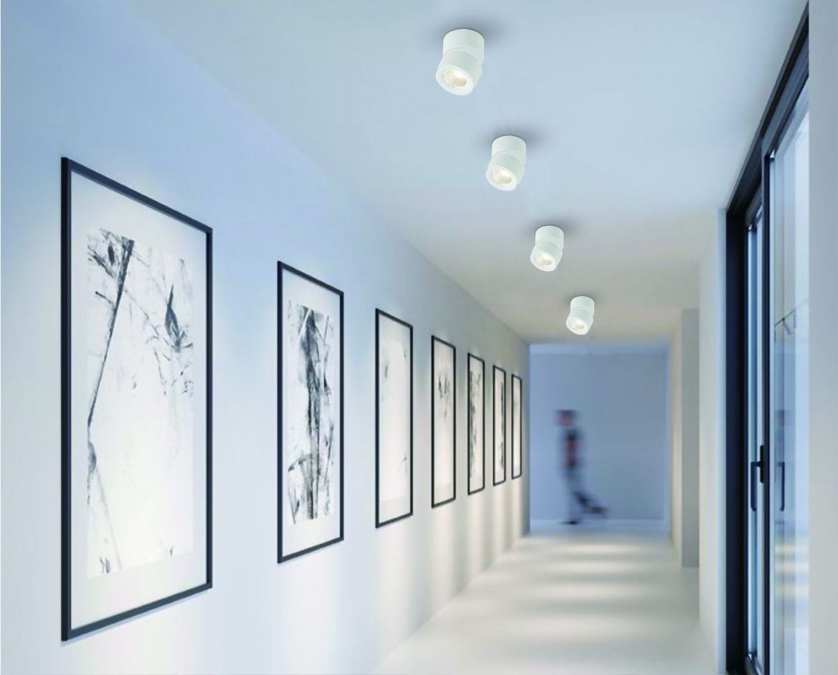 Mone Bianco LED lampa sufitowa 1-punktowa w białym kolorze, zamontowana na suficie w nowoczesnym korytarzu. Oświetlenie skierowane na biało-czarne obrazy w ramkach na ścianie. Prosty, cylindryczny kształt lampy.