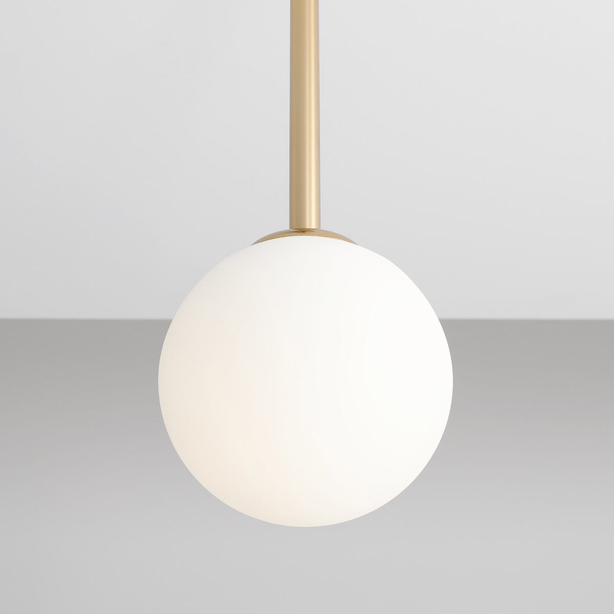 Lampa wisząca Pinne Short 1-punktowa w złotym kolorze, z białą, matową kulą. Ujęcie z bliska, ukazujące detal montażu. Prosta forma, zawieszona na cienkim przewodzie, na szarym tle. Idealna do nowoczesnych wnętrz.