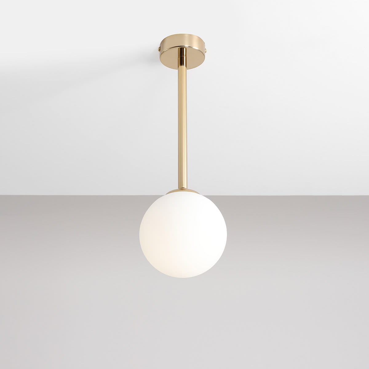 Lampa wisząca Pinne Short w złotym kolorze, z białym, okrągłym kloszem. Zamontowana na długim, złotym stelażu, wisi z sufitu na tle szarej ściany. Minimalistyczny design, idealna do nowoczesnych wnętrz.