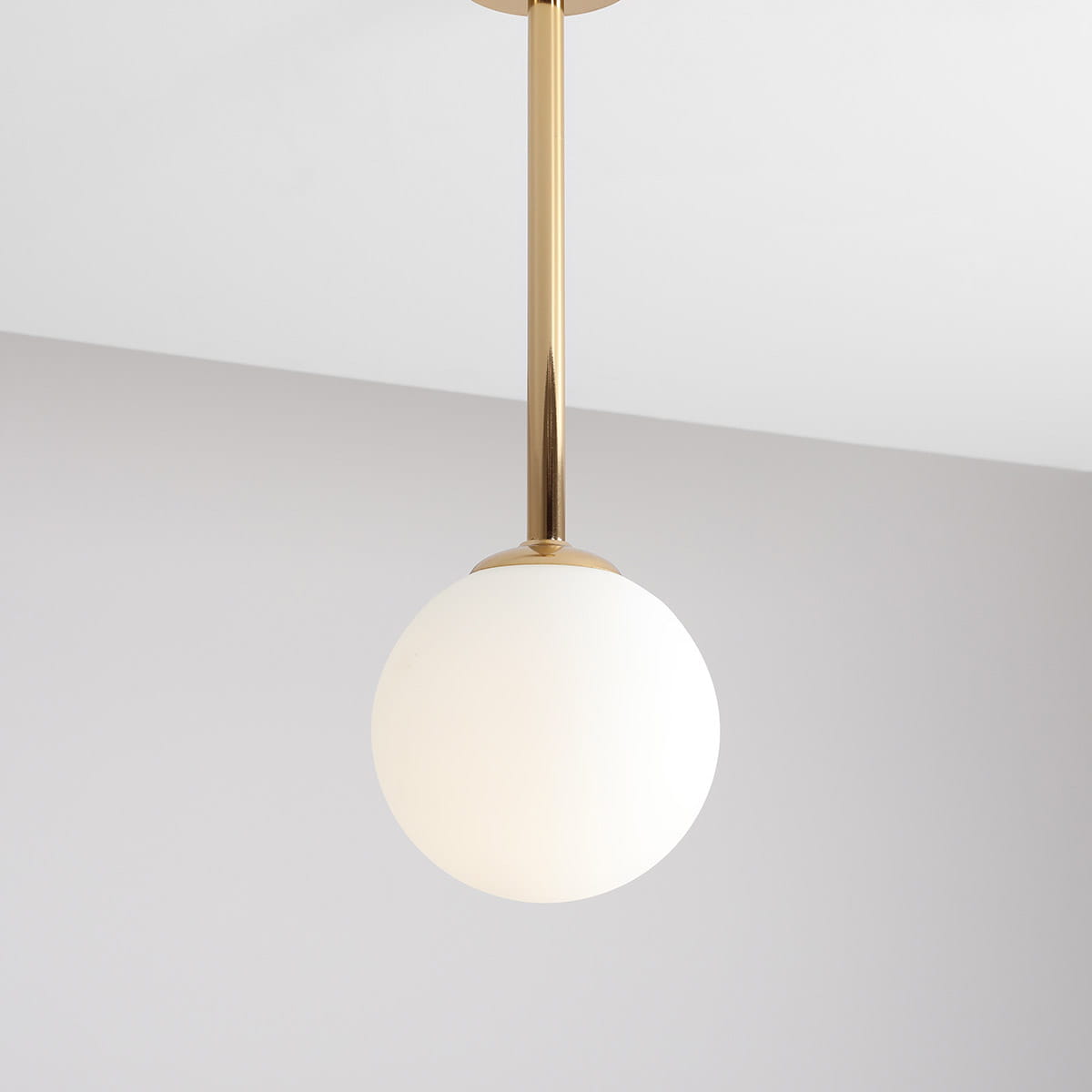 Lampa wisząca Pinne Short w złotym kolorze z białym, okrągłym kloszem, zawieszona na cienkiej, metalowej rurce. Ujęcie z góry, pokazujące elegancki design i gładką fakturę. Tło w jasnym odcieniu, co podkreśla wygląd lampy.