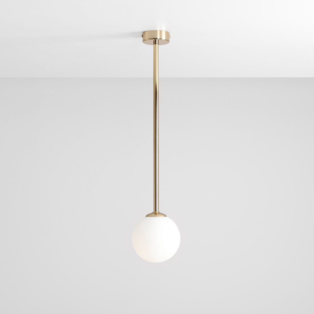 Lampa wisząca Pinne Medium w kolorze złotym, z białym, kulistym kloszem. Zawieszona na długim, prostym stelażu, montowana do sufitu. Tło jasnoszare, co podkreśla elegancki design lampy.
