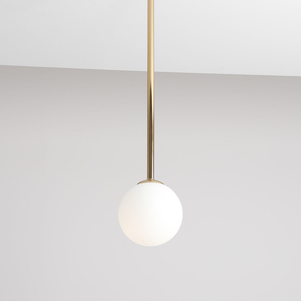 Lampa wisząca Pinne Medium w kolorze złotym, z białym, okrągłym kloszem. Montowana na długim, cienkim stelażu. Klosz wykonany z matowego szkła, zawieszony w centralnej pozycji, na jednolitym, jasnym tle.