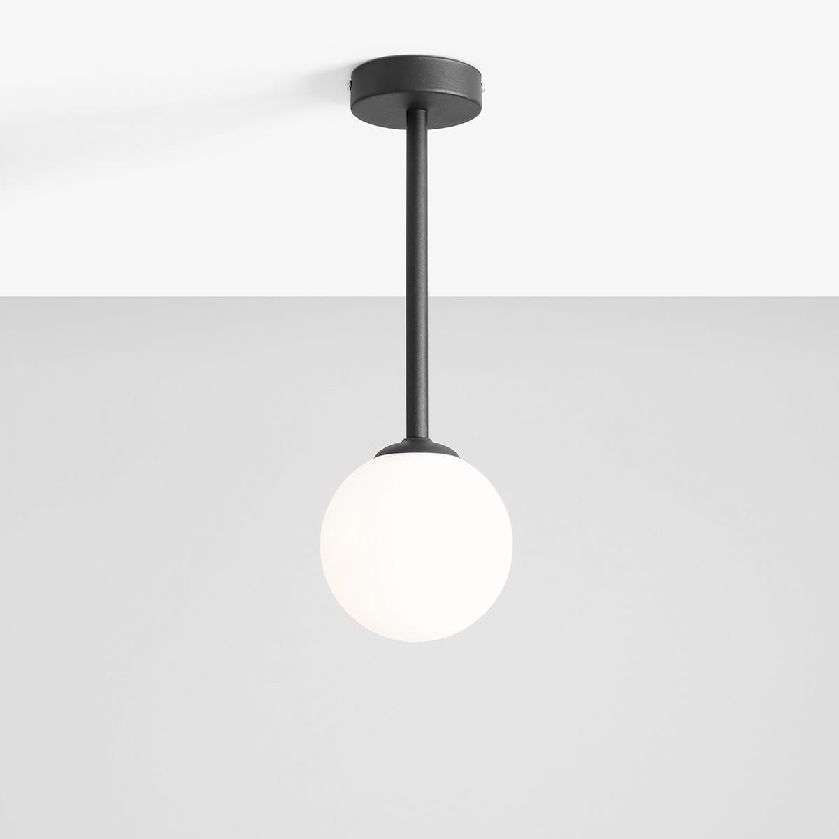 Czarna lampa wisząca Pinne Short z jednym punktem świetlnym, z okrągłym, białym kloszem. Produkt zawieszony na długim, prostym ramieniu, montowany do sufitu. Tło w jasnoszarym kolorze, minimalistyczna aranżacja.