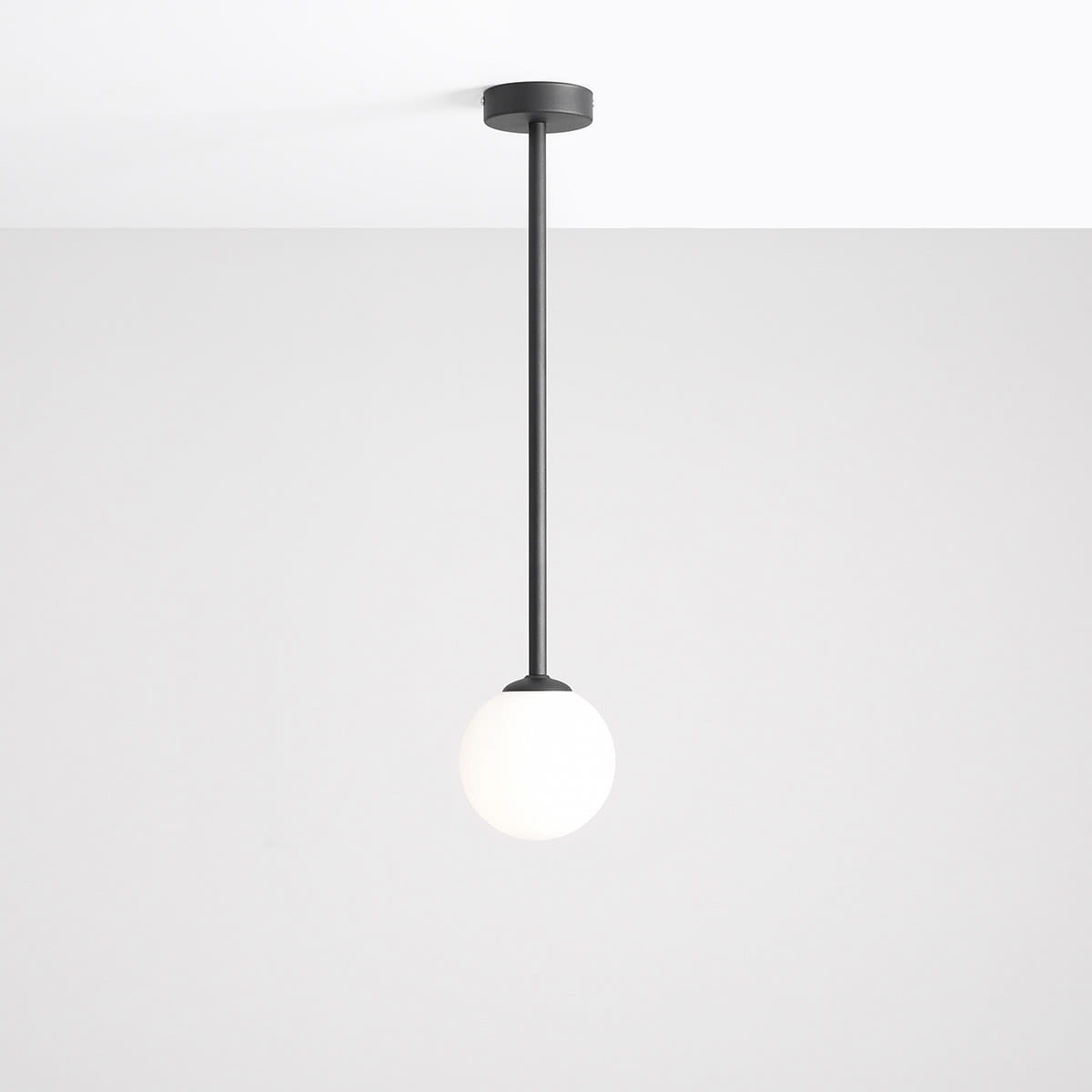 Czarna lampa wisząca Pinne Medium z pojedynczym punktem świetlnym, z długim metalowym ramieniem i okrągłą, matową białą kloszem, zawieszona na prostym, okrągłym mocowaniu sufitowym, na jasnym tle.