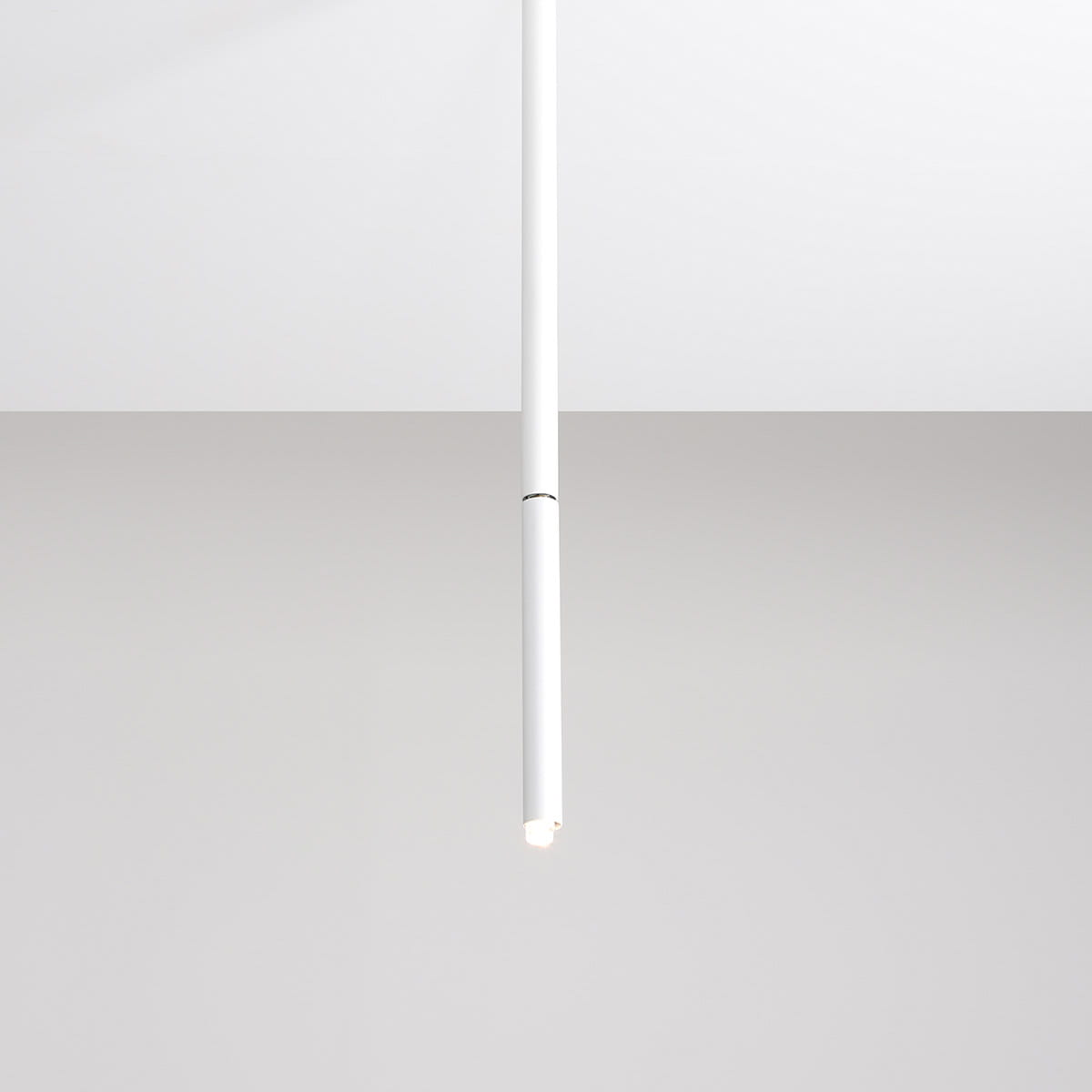 Biała lampa sufitowa Stick M, jednopunktowa, o cylindrycznym kształcie. Zawieszona centralnie, z widocznym źródłem światła na końcu. Tło jasne, minimalistyczne, podkreślające nowoczesny design lampy.