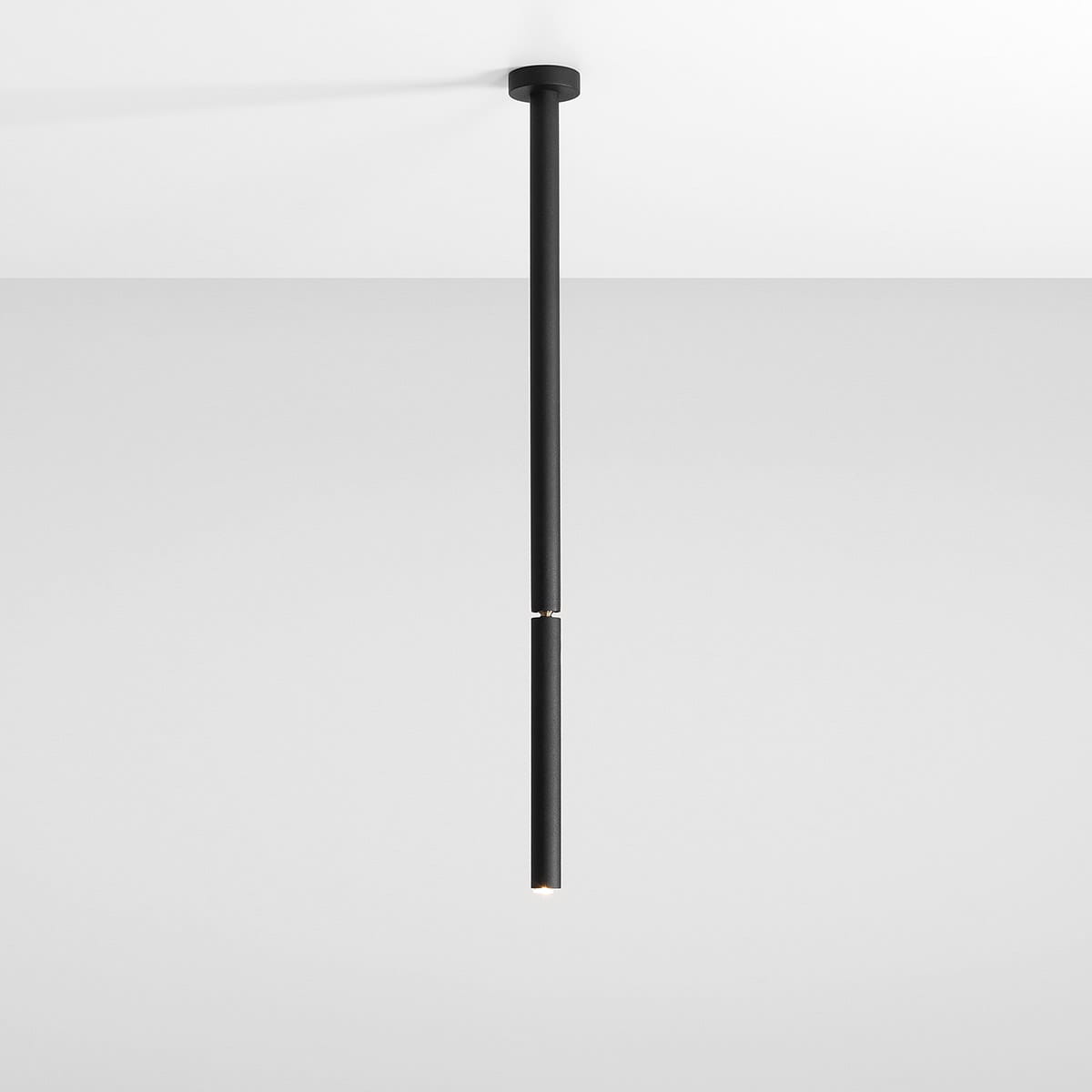 Czarna lampa sufitowa Stick M w formie jednopunktowej. Długa, cylindryczna konstrukcja z matowego materiału, zawieszona na prostym uchwycie. Widoczna jasna żarówka na końcu lampy, umieszczona w minimalistycznym otworze. Tło jasne, jednolite.