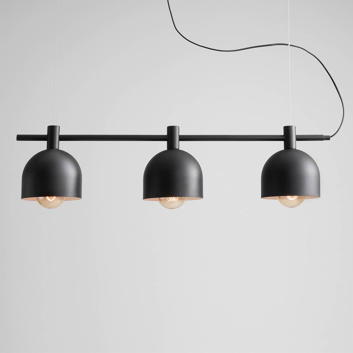 Czarna lampa wisząca Beryl z trzema punktami świetlnymi. Metalowe klosze o kształcie półkulistym, zawieszone na prostym, poziomym wsporniku. Lampa zawieszona na cienkim przewodzie na jasnym tle.