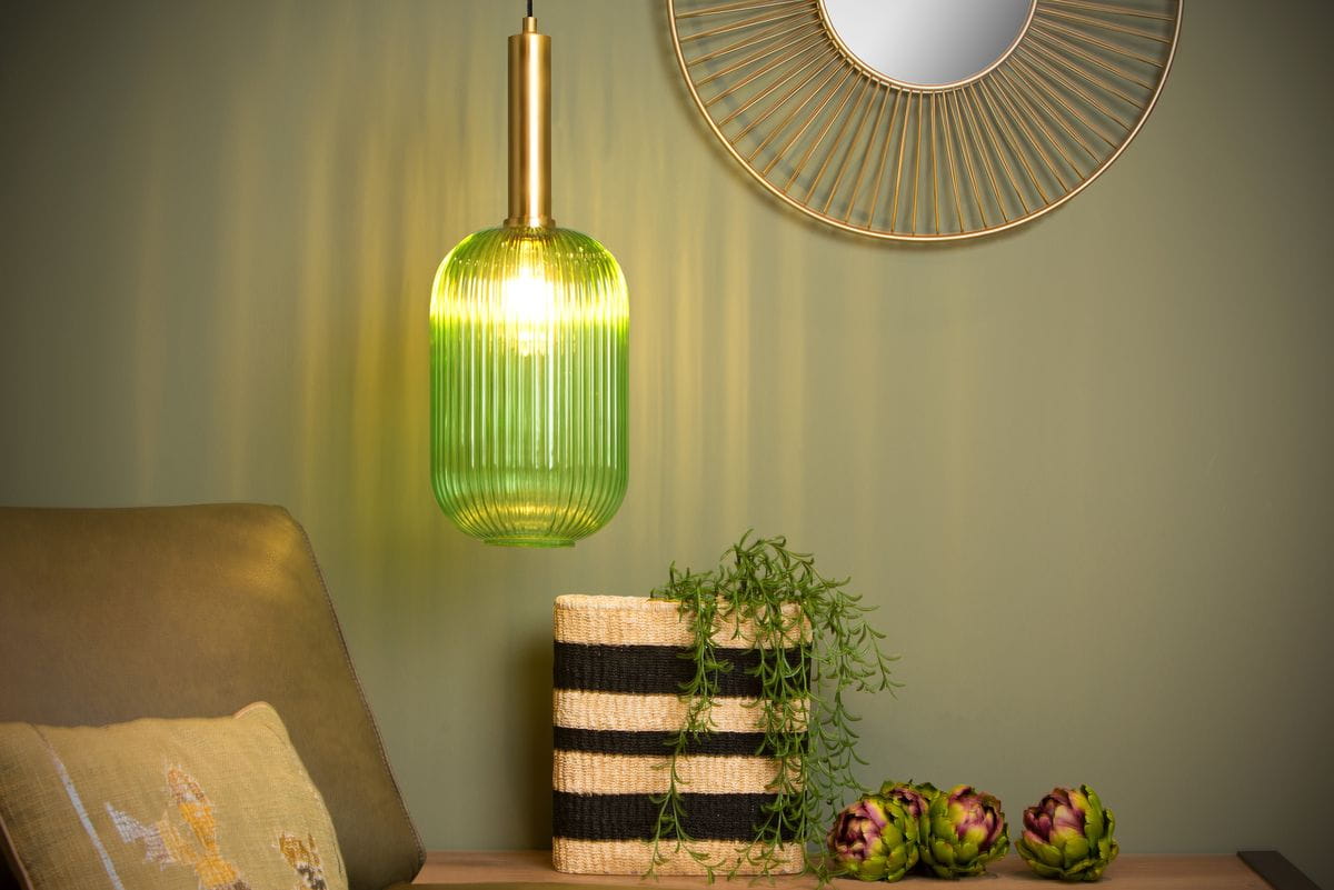 Lampa wisząca 1-punktowa w kolorze zielonym z mosiężnym mocowaniem, o cylindrycznym kształcie z pionowymi rowkami. Zawieszona nad sofą, w tle ściana w odcieniu zieleni oraz lustro w złotej ramie. Widać również kosz z rośliną i dekoracyjne elementy.