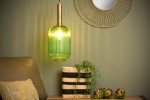 Lampa wisząca 1-punktowa w kolorze zielonym z mosiężnym mocowaniem, o cylindrycznym kształcie z pionowymi rowkami. Zawieszona nad sofą, w tle ściana w odcieniu zieleni oraz lustro w złotej ramie. Widać również kosz z rośliną i dekoracyjne elementy.