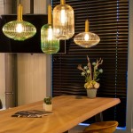 Lampa wisząca Maloto 1-punktowa w kolorze zielonym i mosiądzu, z teksturowanym kloszem, zawieszona nad drewnianym stołem. W tle czarne żaluzje oraz dekoracje roślinne. Wyraźne źródło światła, z eleganckim wykończeniem.