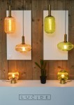 Lampa wisząca Maloto 1-punktowa w kolorze bursztynowym z mosiężnymi detalami. Przezroczysta, teksturowana klosz o cylindrycznym kształcie, zawieszony na czarnym przewodzie. W tle białe panele, drewniana ściana oraz inne lampy w różnych kolorach.