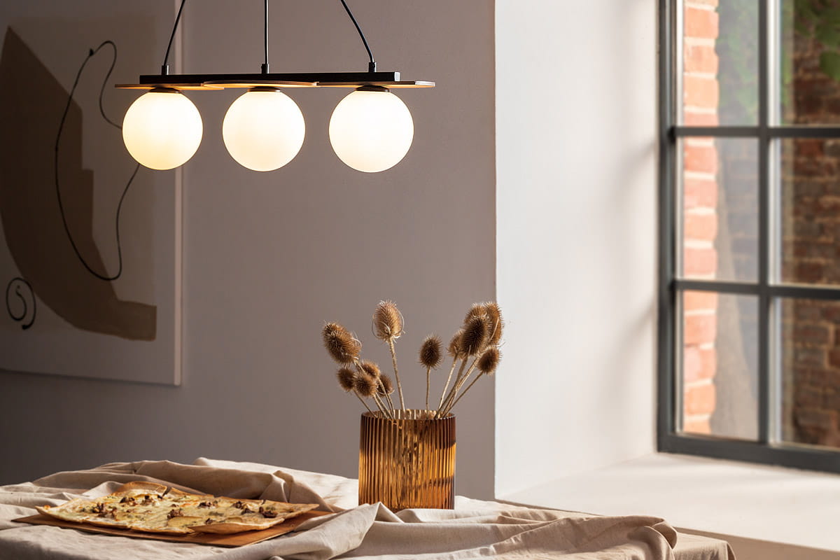 Czarna lampa wisząca Mirror z trzema okrągłymi kloszami w białym kolorze, zawieszona nad stołem. Lampa ma minimalistyczny design z prostą konstrukcją. W tle widoczna jest dekoracja oraz okno, co tworzy przytulną atmosferę.