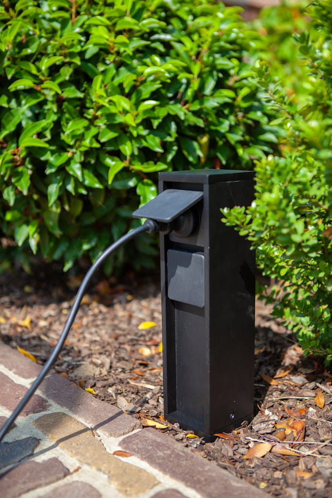 Słupek ogrodowy Mains w kolorze czarnym, z dwoma wbudowanymi gniazdami typu F. Ujęcie z boku, gniazdo z kablem włożonym, umieszczony na podłożu z korą, w tle zieleń krzewów.
