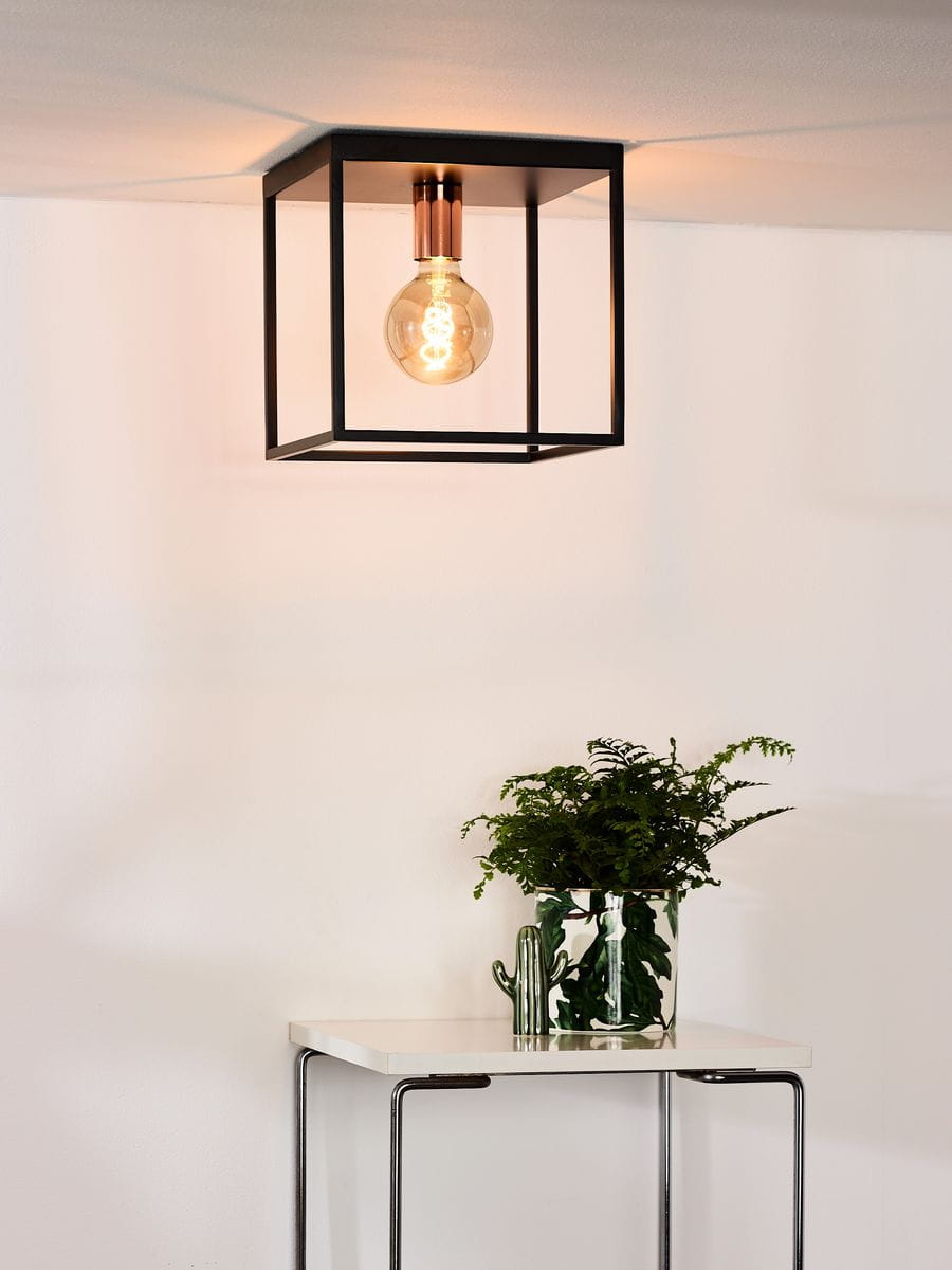 Czarna lampa sufitowa Arthur 1-punktowa z metalową ramą w kształcie sześcianu, z przezroczystą żarówką LED. Montowana na suficie, w otoczeniu białej ściany i nowoczesnego stolika z roślinnością.