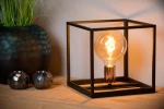 Czarna lampa stołowa Arthur 1-punktowa w metalowej ramie, z okrągłą żarówką w stylu retro, umieszczona na drewnianym stole. W tle widoczna dekoracyjna doniczka oraz dwa metalowe świeczniki. Całość oświetlona ciepłym światłem.