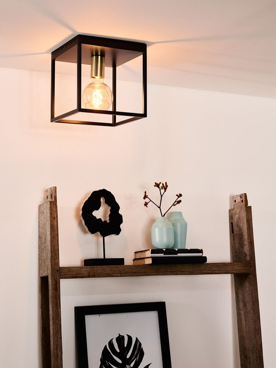 Czarna lampa sufitowa Ruben 1-punktowa o kwadratowej formie, z widoczną żarówką. Metalowa konstrukcja z mosiężnym elementem na górze. Lampa zamontowana na białym tle, nad drewnianą półką z dekoracjami.