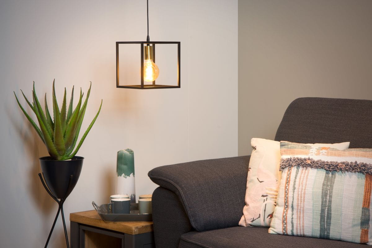 Czarna lampa wisząca Ruben 1-punktowa w formie prostej metalowej konstrukcji z przezroczystą żarówką, zawieszona nad szarym fotelem. W tle widać zieloną roślinę w czarnym doniczce oraz dekoracyjne elementy na stoliku.