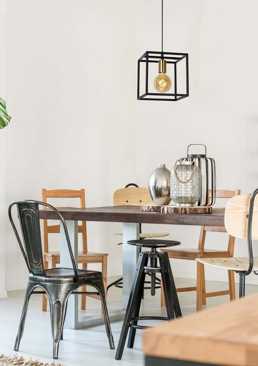 Czarna lampa wisząca Ruben 1-punktowa o geometrycznym kształcie, zawieszona nad drewnianym stołem. Wewnątrz przezroczysta żarówka. W tle widać różne krzesła i dekoracyjne akcesoria. Tło jasne, minimalistyczne.