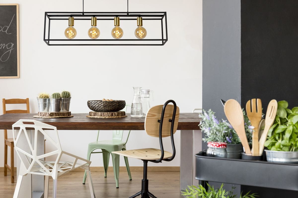 Czarna lampa wisząca Ruben 4-punktowa z przezroczystymi żarówkami, zawieszona nad drewnianym stołem w nowoczesnej jadalni. Widoczna metalowa konstrukcja w formie prostokąta. W tle rośliny i różnorodne krzesła.