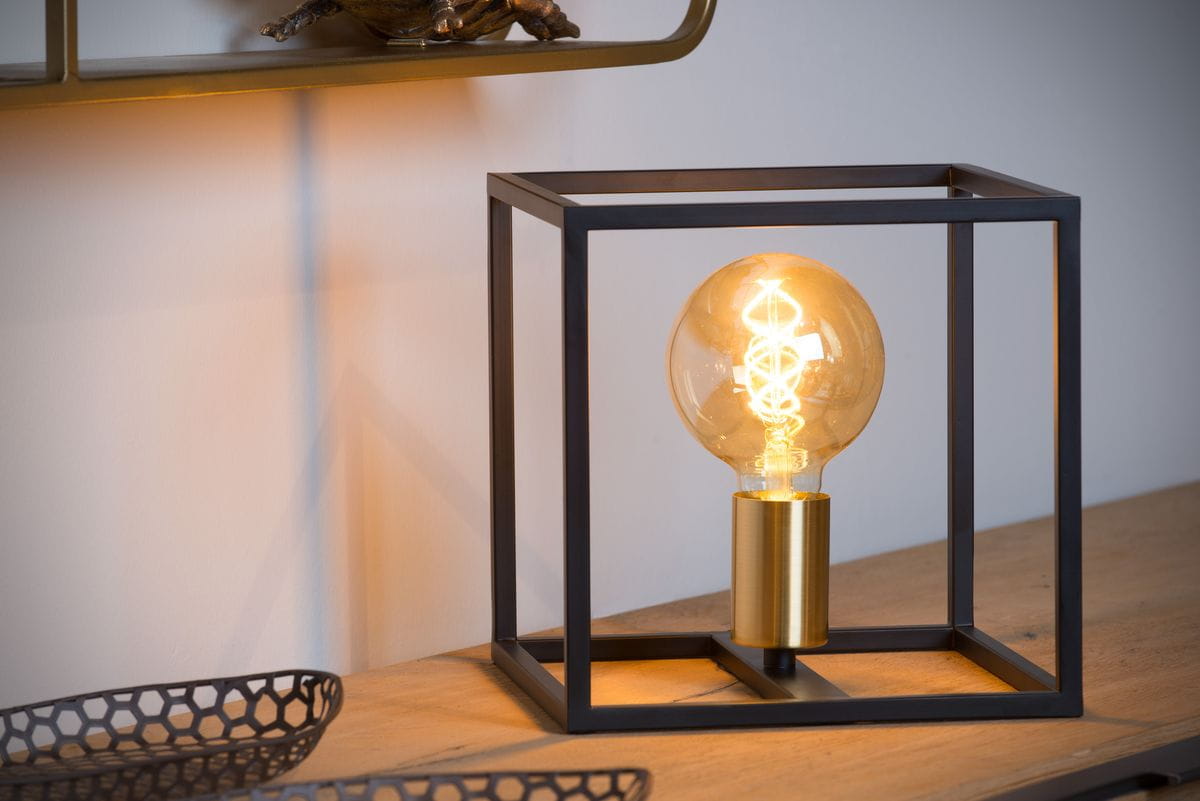 Czarna lampa stołowa Ruben 1-punktowa z metalową ramą w kształcie sześcianu. Wewnątrz znajduje się żarówka w stylu retro z mosiężnym mocowaniem, emitująca ciepłe światło. Lampa ustawiona na drewnianym blacie, w tle jasna ściana.