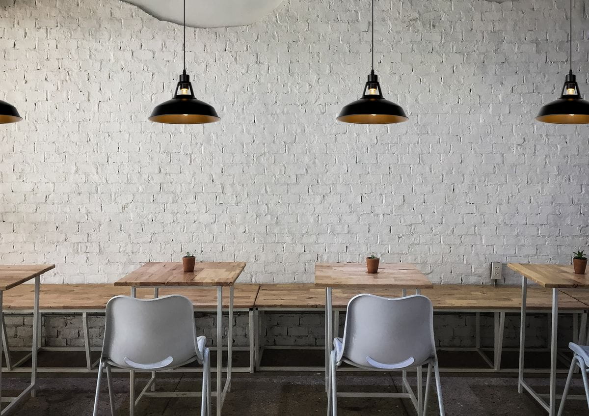 Czarna lampa wisząca Brassy-Bis 1-punktowa z metalowym kloszem w industrialnym stylu, zawieszona nad drewnianymi stołami w jasnym wnętrzu z białą ceglaną ścianą. W tle widoczne białe krzesła i małe rośliny w doniczkach.