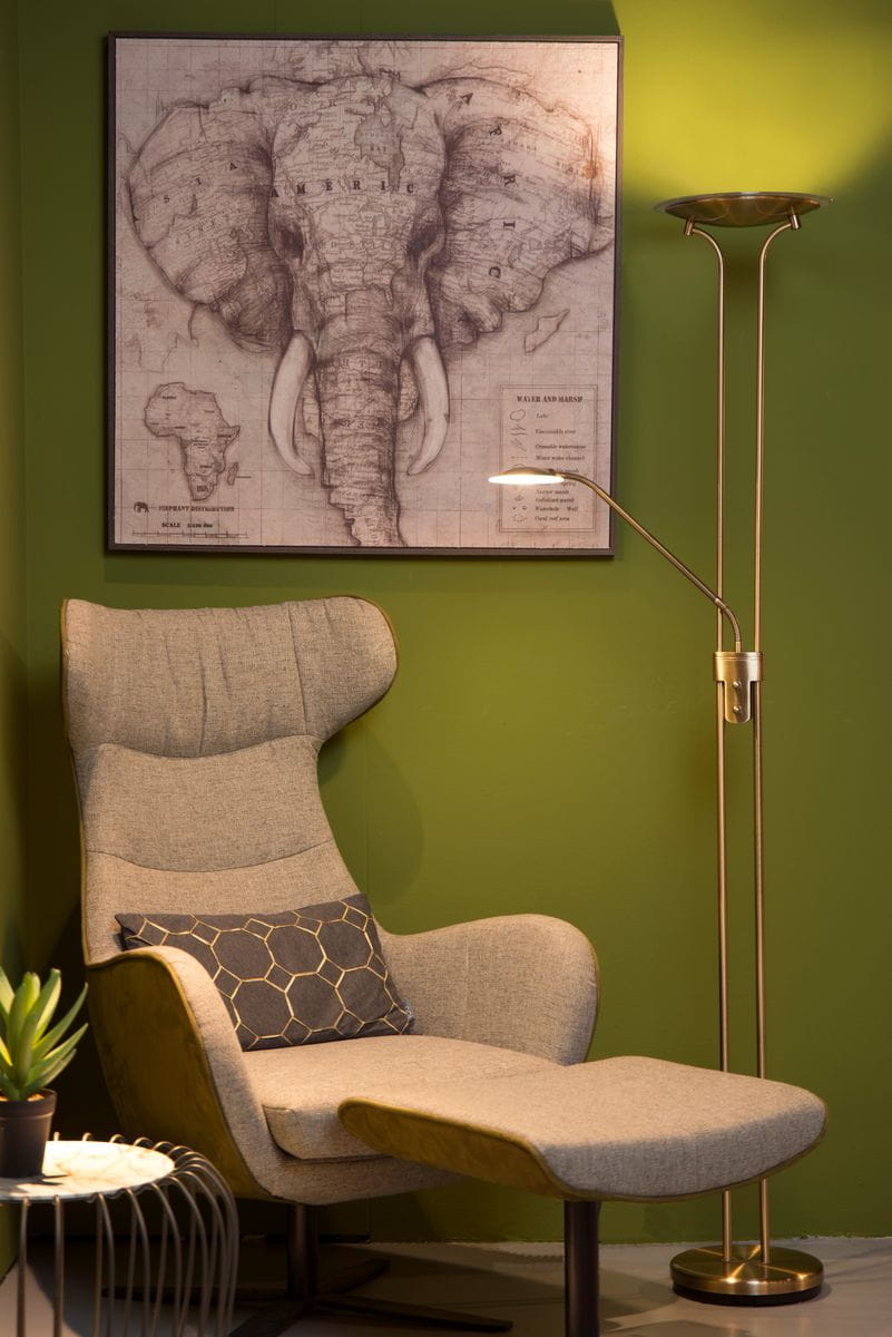Lampa podłogowa Champion LED, 2-punktowa, w kolorze antycznego mosiądzu. Ustawiona w narożniku, z długą, smukłą nogą i okrągłą podstawą. Obok wygodny fotel z podnóżkiem, w tle zielona ściana oraz obraz przedstawiający słonia.