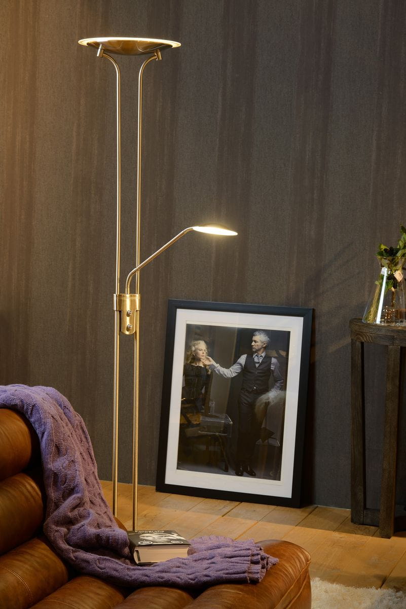 Lampa podłogowa Champion LED 2-punktowa w satynowym chromie, z jedną dużą górną lampą i regulowaną lampą boczną. Stoi na stabilnej podstawie, obok skórzanej kanapy, na której leży fioletowy koc. W tle zdjęcie i drewniany stolik.