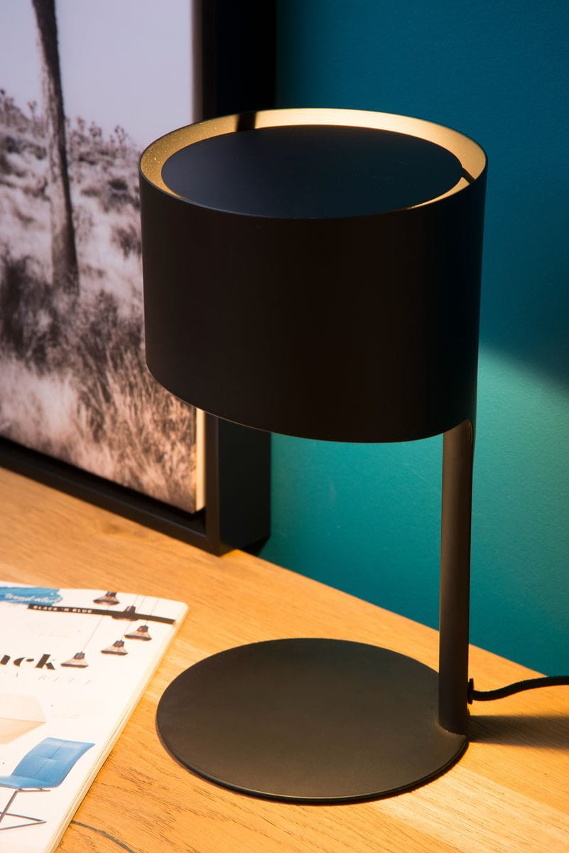 Czarna lampa stołowa Knulle 1-punktowa, z cylindrycznym kloszem i okrągłą podstawą. Ujęcie z boku, ukazujące minimalistyczny design i złote akcenty w górnej części klosza. Lampa znajduje się na drewnianym blacie, obok magazynu.