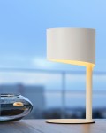 Lampa stołowa Knulle 1-punktowa w kolorze białym, z cylindrycznym kloszem i minimalistyczną podstawą. Ujęcie z boku, ukazujące subtelne podświetlenie. Produkt umieszczony na drewnianym stole, w tle widoczny niebieski horyzont.