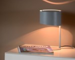 Lampa stołowa Knulle 1-punktowa w kolorze szarym, z okrągłym kloszem i smukłą podstawą. Ujęcie z boku, lampa emituje ciepłe światło, a obok znajduje się magazyn na białym stole. Tło w neutralnym odcieniu, podkreślającym elegancję lampy.