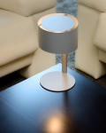 Lampa stołowa Knulle 1-punktowa w kolorze szarym z okrągłym kloszem i metalową podstawą. Ustawiona na czarnym blacie, w otoczeniu jasnych kanap. Klosz ma złoty akcent na górze, co podkreśla nowoczesny design.