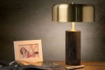 Lampa stołowa Mirasol 3-punktowa w kolorze czarnym, z cylindryczną podstawą i metalowym kloszem w odcieniu złota. Ujęcie z boku, na drewnianym biurku, obok zdjęcie w ramce i magazyn. Klosz tworzy ciepłe światło, podkreślając elegancki design.