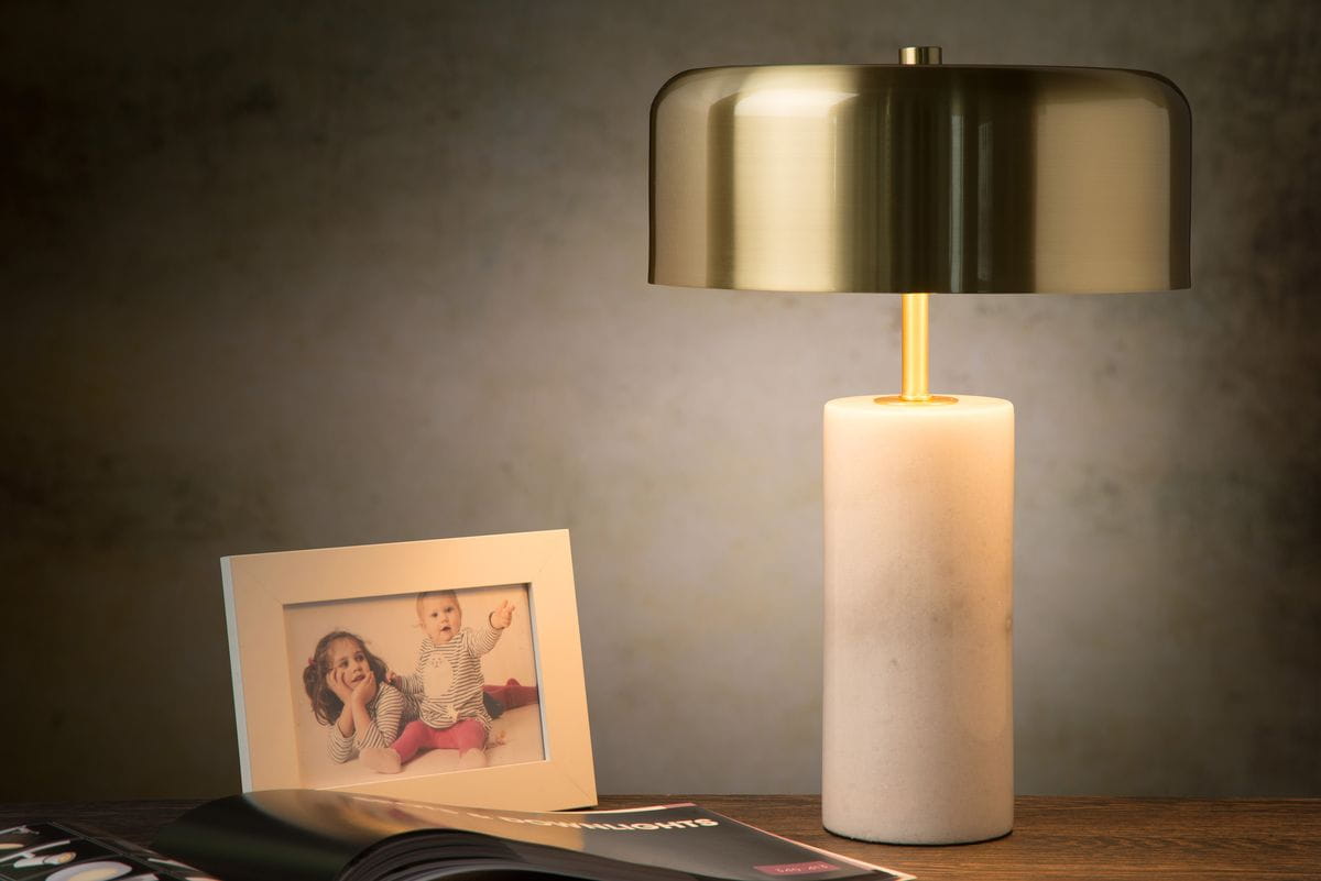 Lampa stołowa Mirasol 3-punktowa w białej obudowie z okrągłym kloszem w kolorze złotym. Ustawiona na drewnianym stole obok ramki ze zdjęciem i magazynem. Klosz ma gładką, błyszczącą powierzchnię, a podstawa jest cylindryczna, matowa.