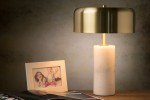 Lampa stołowa Mirasol 3-punktowa w białej obudowie z okrągłym kloszem w kolorze złotym. Ustawiona na drewnianym stole obok ramki ze zdjęciem i magazynem. Klosz ma gładką, błyszczącą powierzchnię, a podstawa jest cylindryczna, matowa.