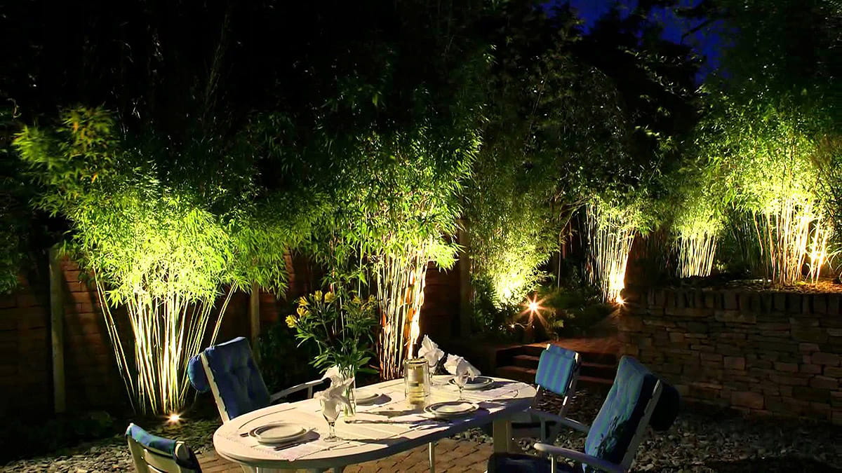 Czarna oprawa dogruntowa Grassi na szpikulcu, umieszczona w ogrodzie, oświetlająca roślinność. Produkt stoi w ziemi, tworząc efektowne iluminacje wokół, na tle ciemnego otoczenia. Szpikulce pozwalają na łatwe umiejscowienie w różnych lokalizacjach.