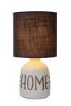Lampa stołowa Cosby 1-punktowa z białą, ceramiczną podstawą z napisem HOME oraz ciemnobrązowym, tekstylnym kloszem. Ujęcie frontalne, lampa stoi na płaskiej powierzchni, emitując ciepłe światło.