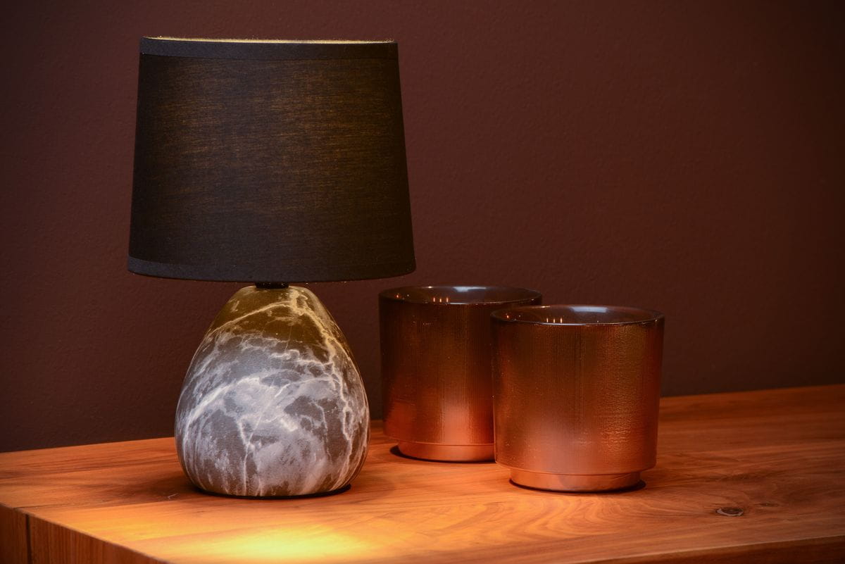 Czarna lampa stołowa Marmo z kamienną podstawą w kształcie łezki i tekstylnym kloszem. Ustawiona na drewnianym blacie obok dwóch metalowych kubków w odcieniu miedzi. Ujęcie z boku, eksponujące detale lampy i otoczenie.