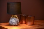 Czarna lampa stołowa Marmo z kamienną podstawą w kształcie łezki i tekstylnym kloszem. Ustawiona na drewnianym blacie obok dwóch metalowych kubków w odcieniu miedzi. Ujęcie z boku, eksponujące detale lampy i otoczenie.