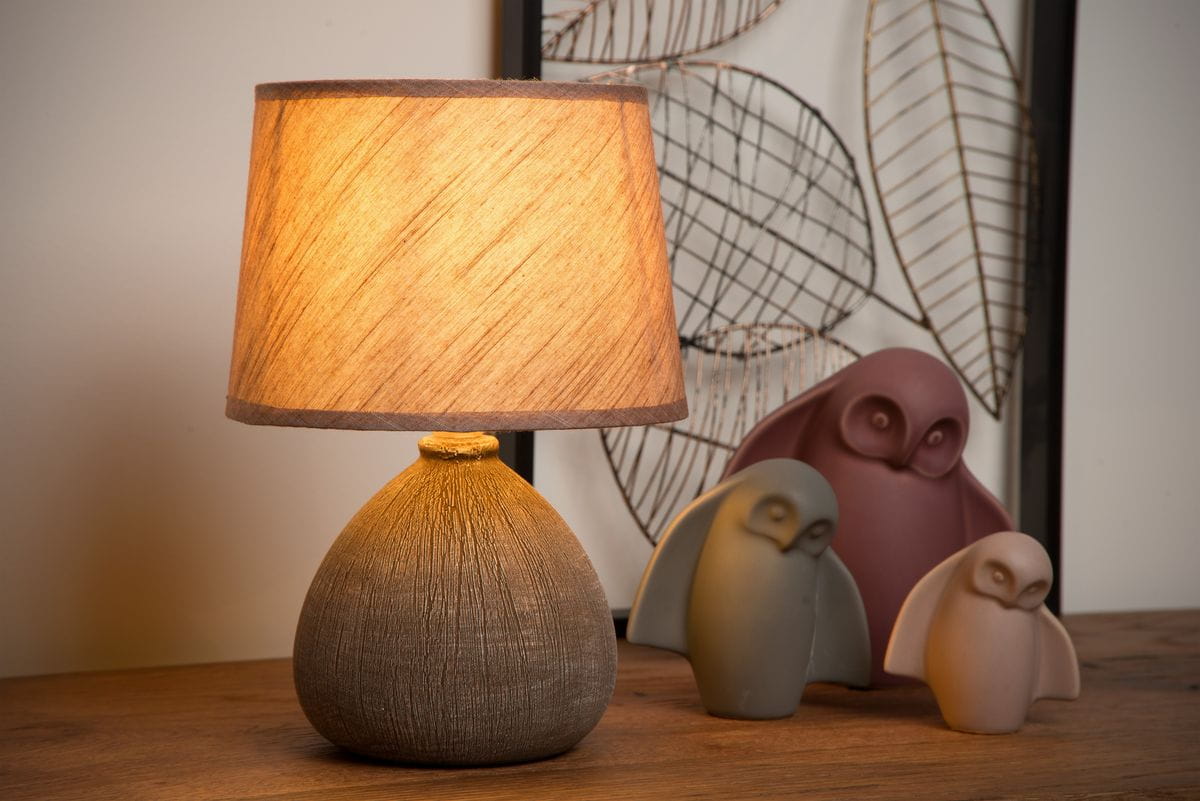 Brązowa lampa stołowa Ramzi z okrągłym, teksturowanym podstawą i kloszem w odcieniu jasnego beżu. Ujęcie frontalne, lampa świeci ciepłym światłem, w tle widoczne dekoracyjne figurki ptaków na drewnianym blacie.
